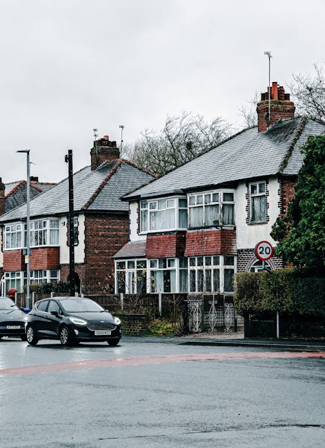 A row of traditional Victorian-style terraced houses constructed from grey stone with white framing around the windows and doors, situated behind a well-maintained grassy park area. Several leafless t